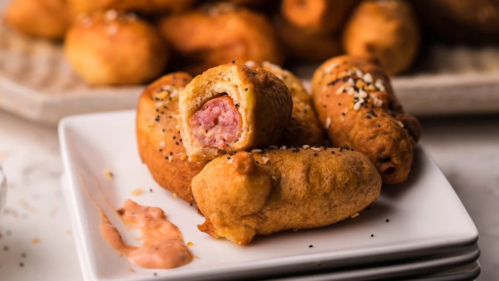 A close-up of several mini corn dogs on a white plate, one with a bite revealing sausage inside, with sauce on the side.
