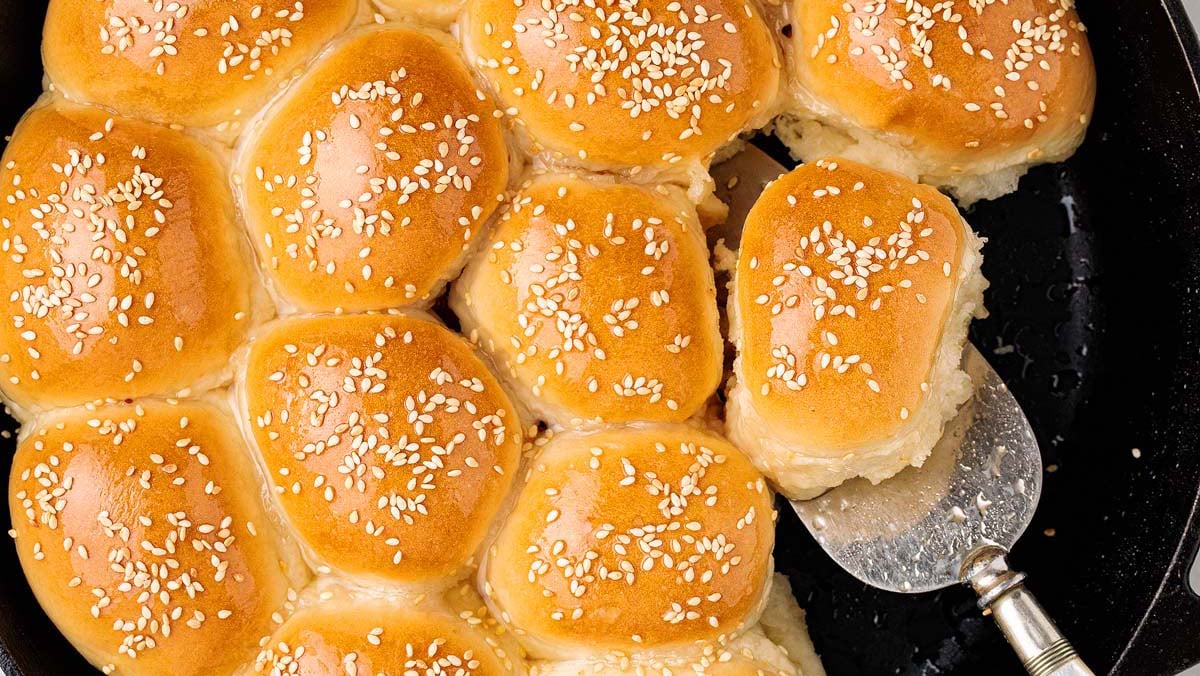A tray of golden brown dinner rolls topped with sesame seeds, with one roll being lifted out using a metal spatula.