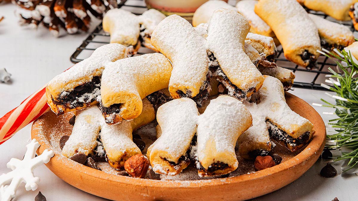 A plate of powdered sugar-covered cookies filled with a dark fruit and nut mixture, with more cookies on a wire rack in the background.