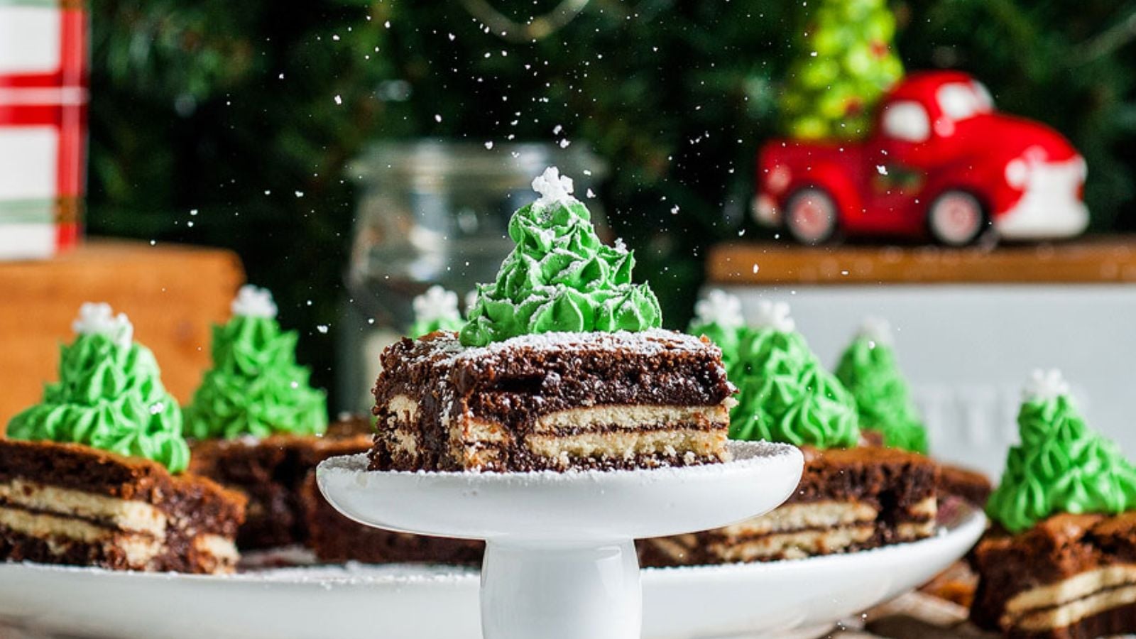 A slice of chocolate layered cake topped with green frosting shaped like a Christmas tree, dusted with powdered sugar, on a white stand. Other decorated slices are in the background.