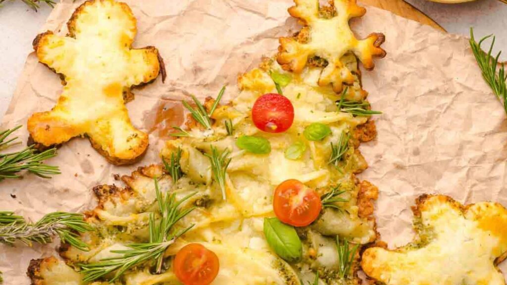 Tree-shaped cheesy flatbread topped with herbs and cherry tomatoes, surrounded by bread cutouts shaped like gingerbread people, on parchment paper.