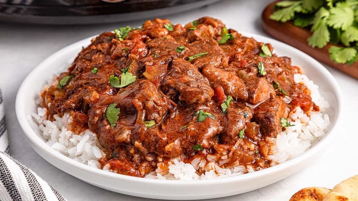 A plate of white rice topped with beef stew in a tomato-based sauce, garnished with chopped cilantro.