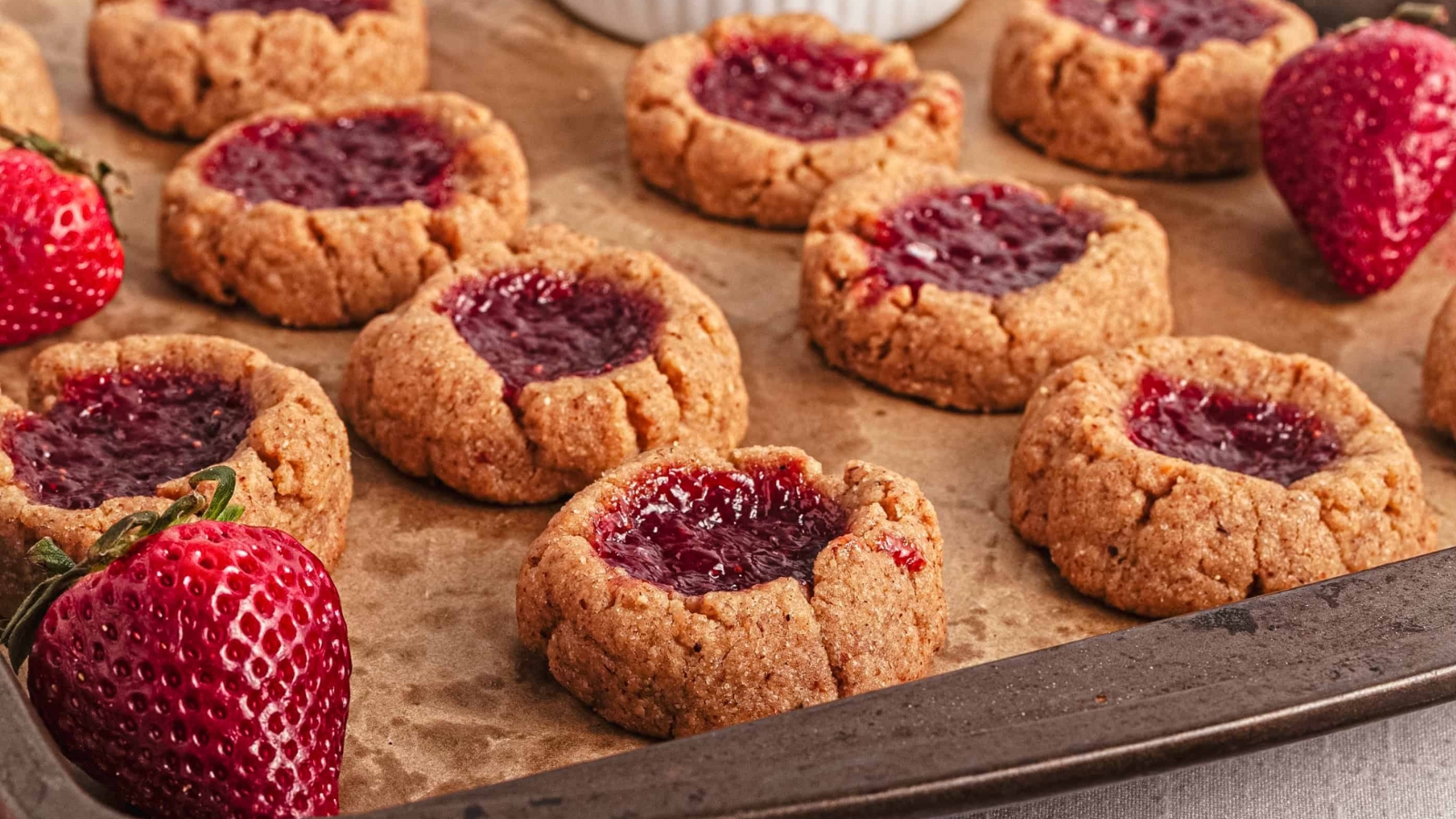 Thumbprint cookies with strawberry jam on a baking tray, surrounded by fresh strawberries.