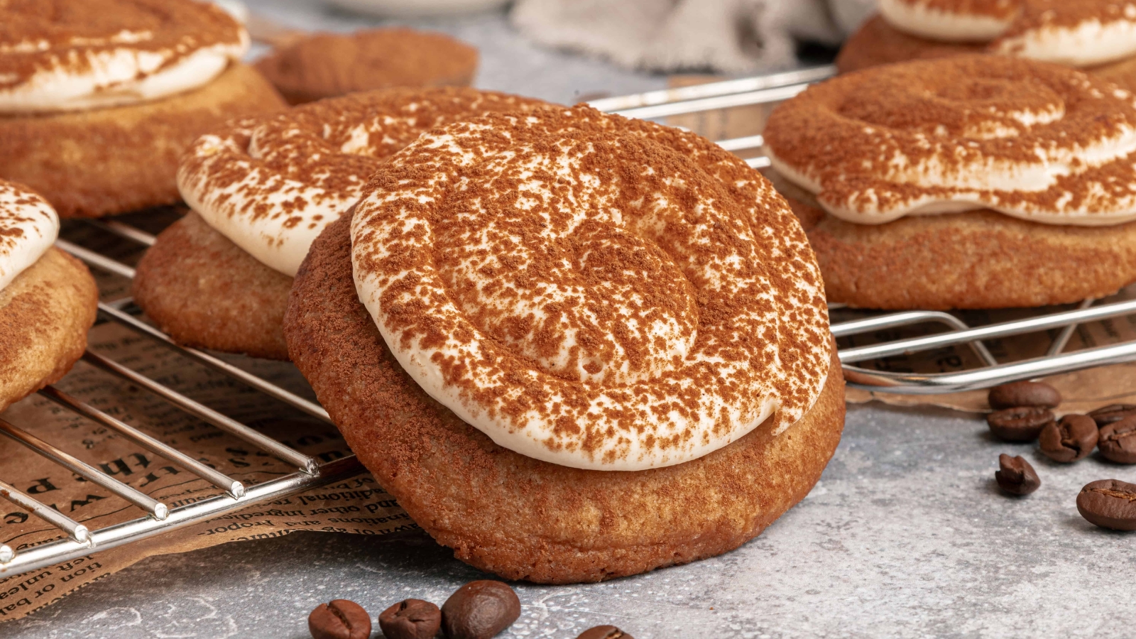 A frosted cookie dusted with cocoa powder, surrounded by coffee beans and baked cookies on a cooling rack.