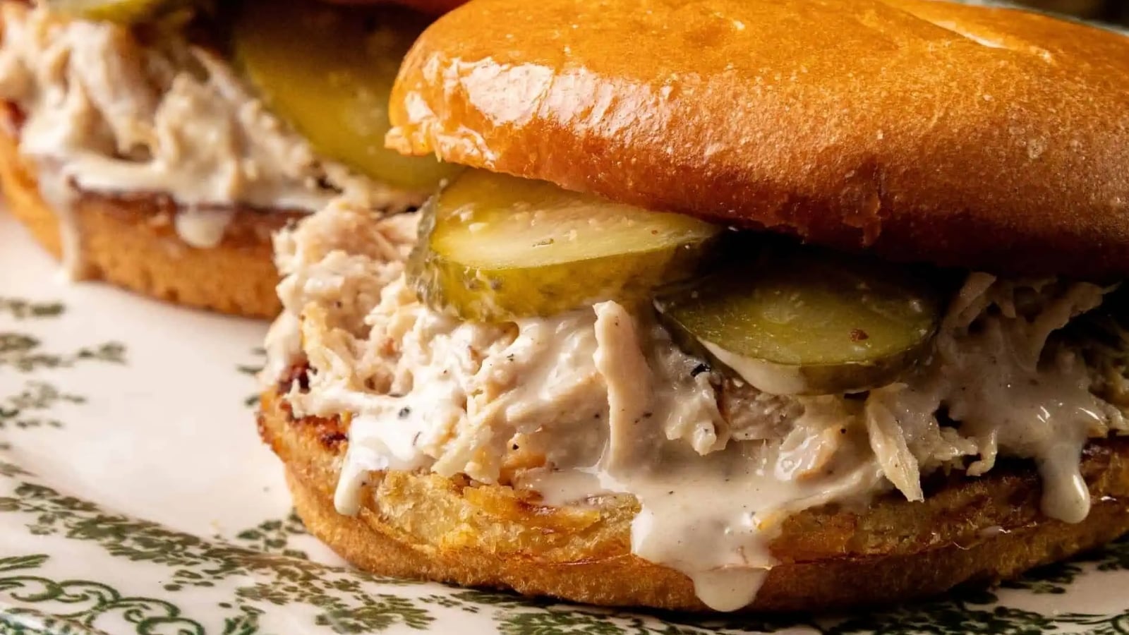 Two Alabama Chopped Chicken Sandwiches with pulled chicken, creamy Alabama white BBQ sauce, and pickle slices on shiny, toasted buns, served on a decorative plate.