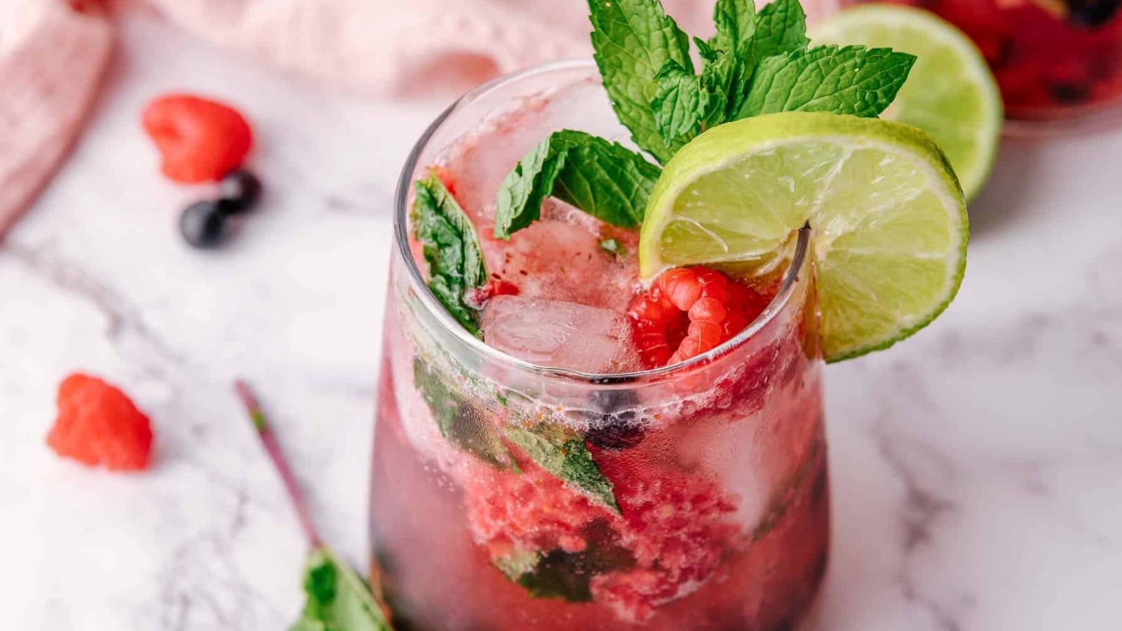 A berry mojito with lime slices, mint leaves, and ice in a glass on a marble surface.