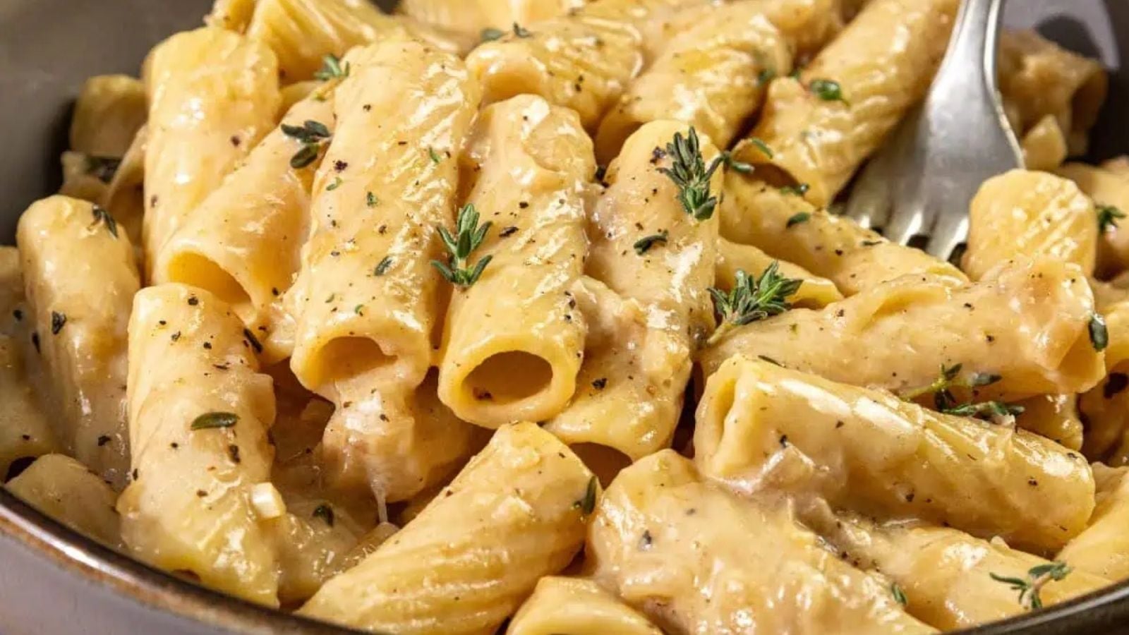 A close-up of creamy French onion pasta rigatoni with caramelized onions, garnished with fresh thyme and cracked black pepper, served in a bowl with a fork.