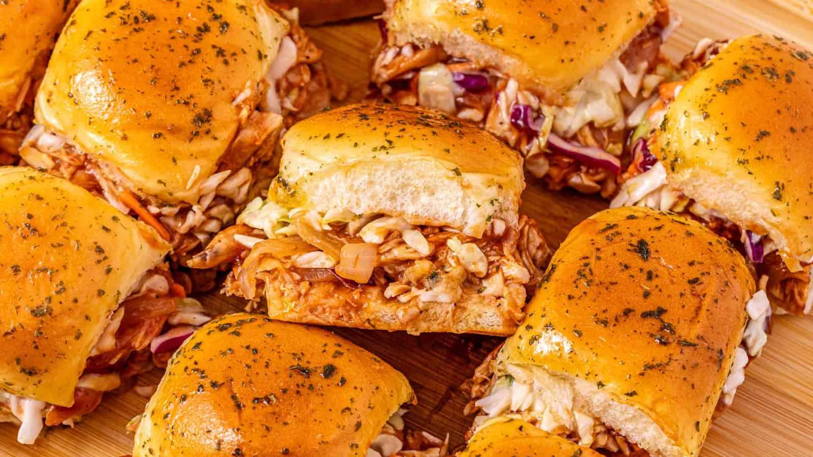 Several barbecue chicken sliders with coleslaw on golden buns are arranged on a wooden surface.