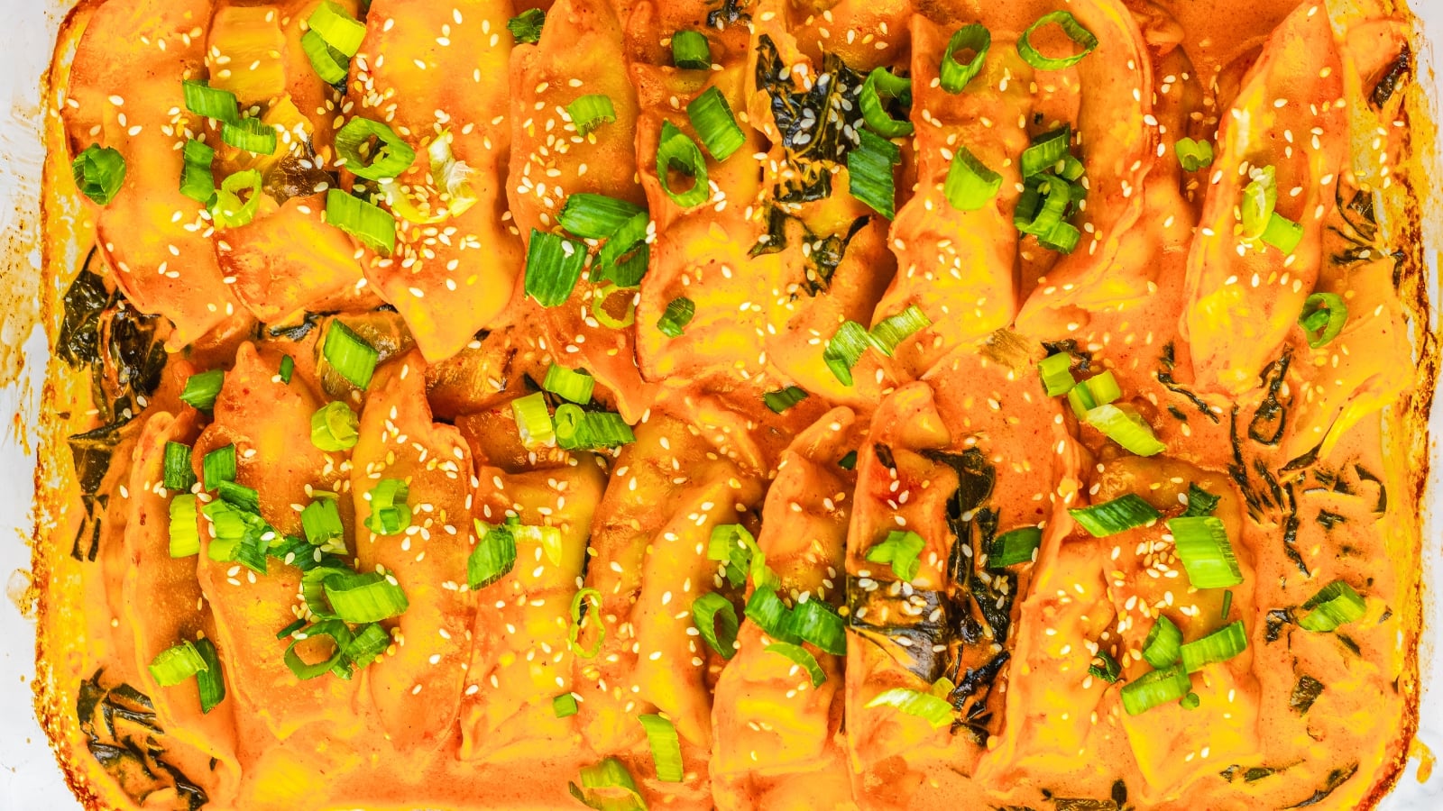 Close-up of baked dumplings in orange sauce, topped with chopped green onions and sesame seeds.