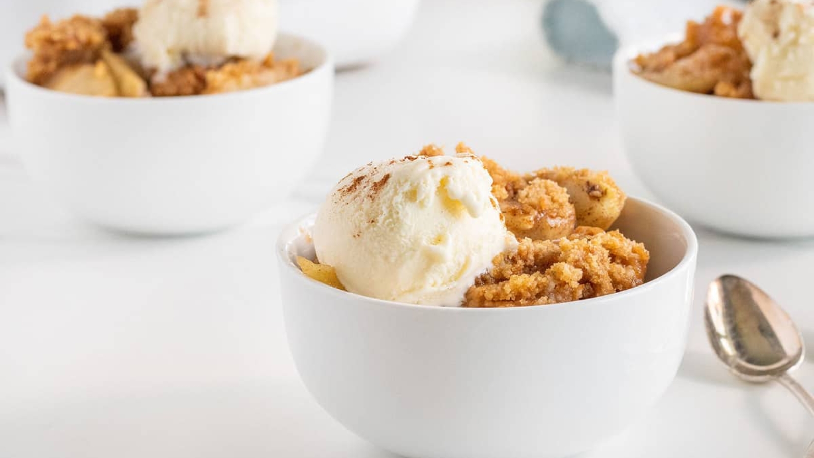 A white bowl filled with apple crisp and a scoop of vanilla ice cream sits on a white surface with a spoon beside it; two more bowls are in the background.