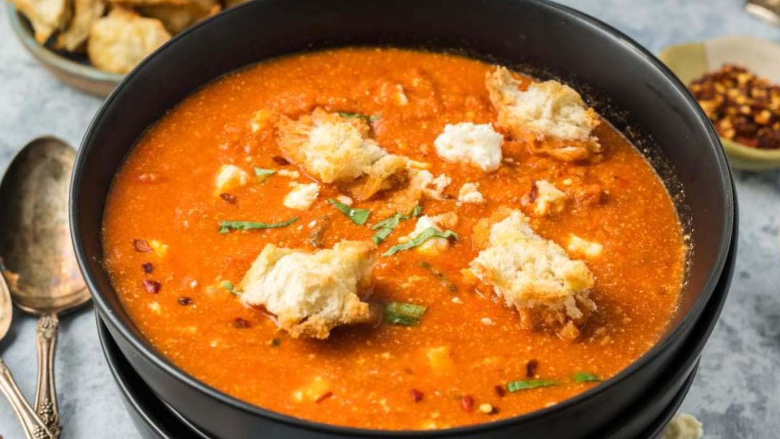 A black bowl filled with tomato soup topped with chunks of bread, cheese, and fresh herbs, next to a spoon and small bowl of red pepper flakes.