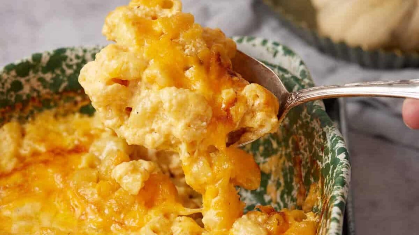 A spoon scoops up cheesy baked macaroni and cheese from a green and white dish.