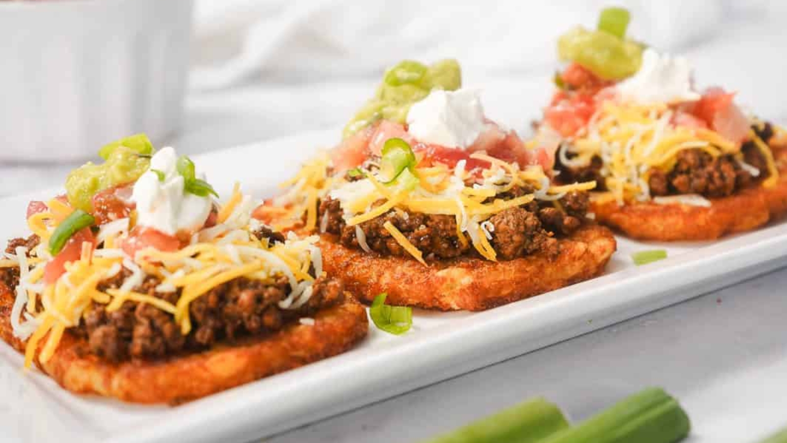 Three hash brown patties topped with ground beef, cheese, sour cream, tomatoes, and green onions on a plate.
