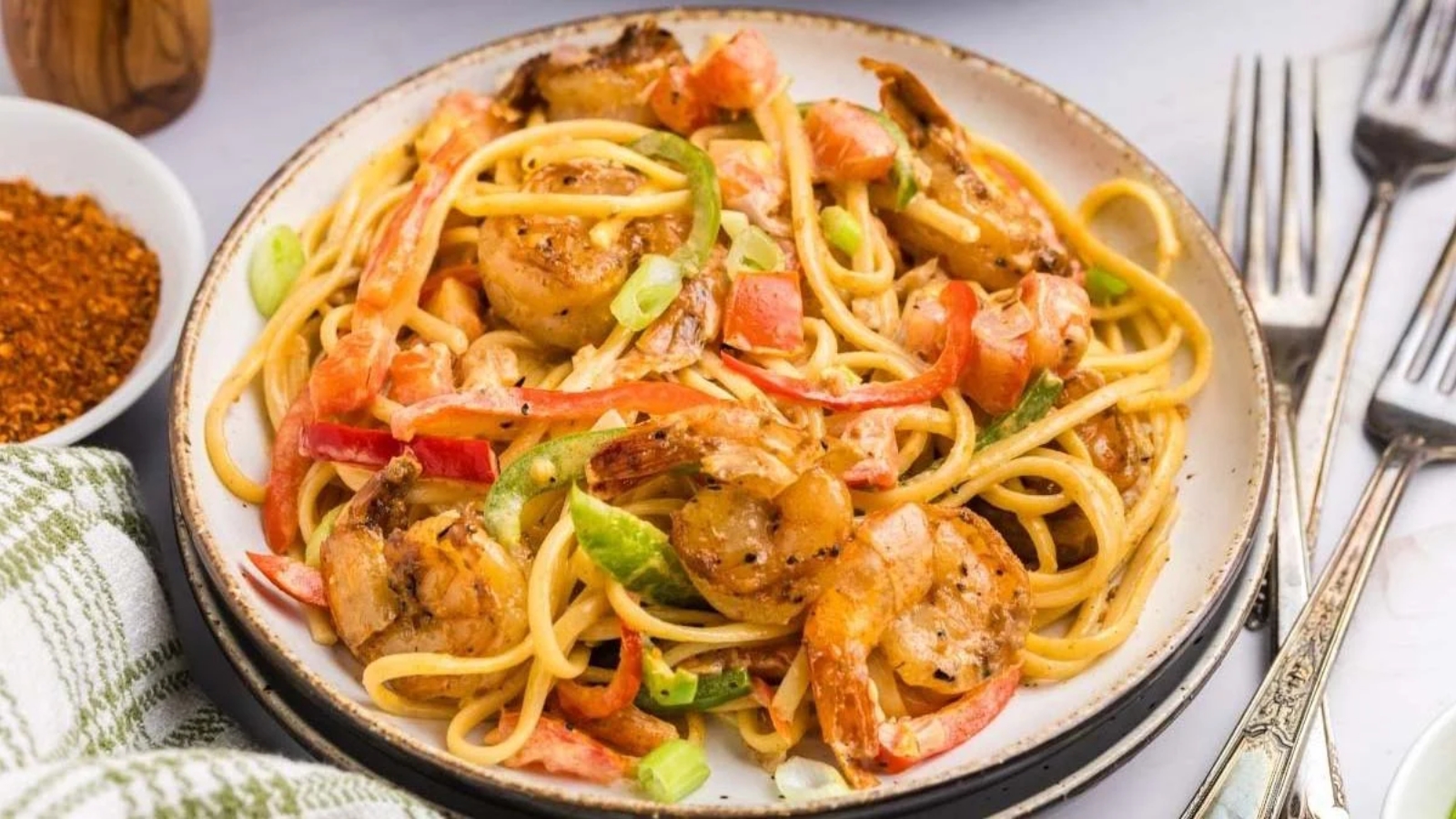 A plate of pasta with shrimp, bell peppers, and green onions, served on a white dish with forks beside it.