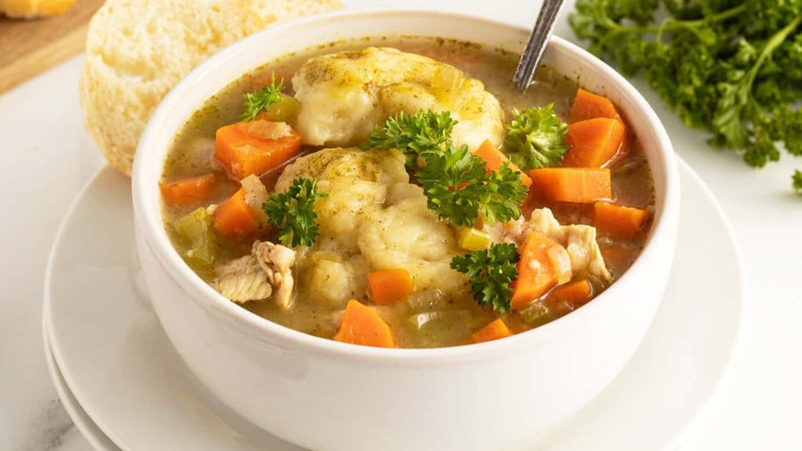 A bowl of chicken and vegetable soup with dumplings, garnished with parsley, served with a piece of bread on a white plate.