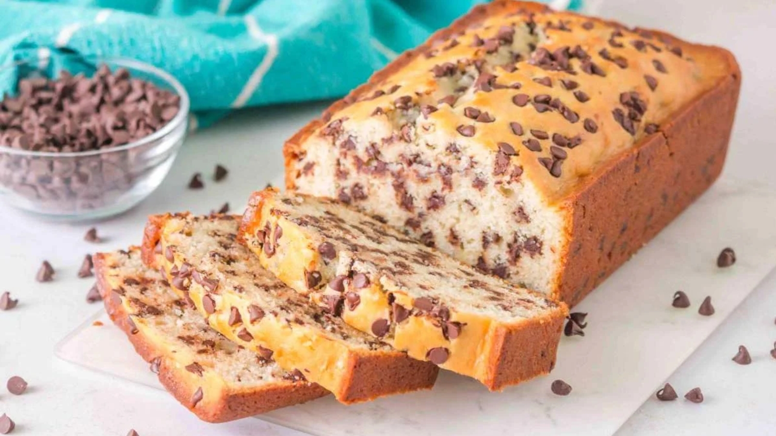 A loaf of chocolate chip bread partially sliced, with chocolate chips scattered on top and around, and a bowl of chocolate chips in the background.