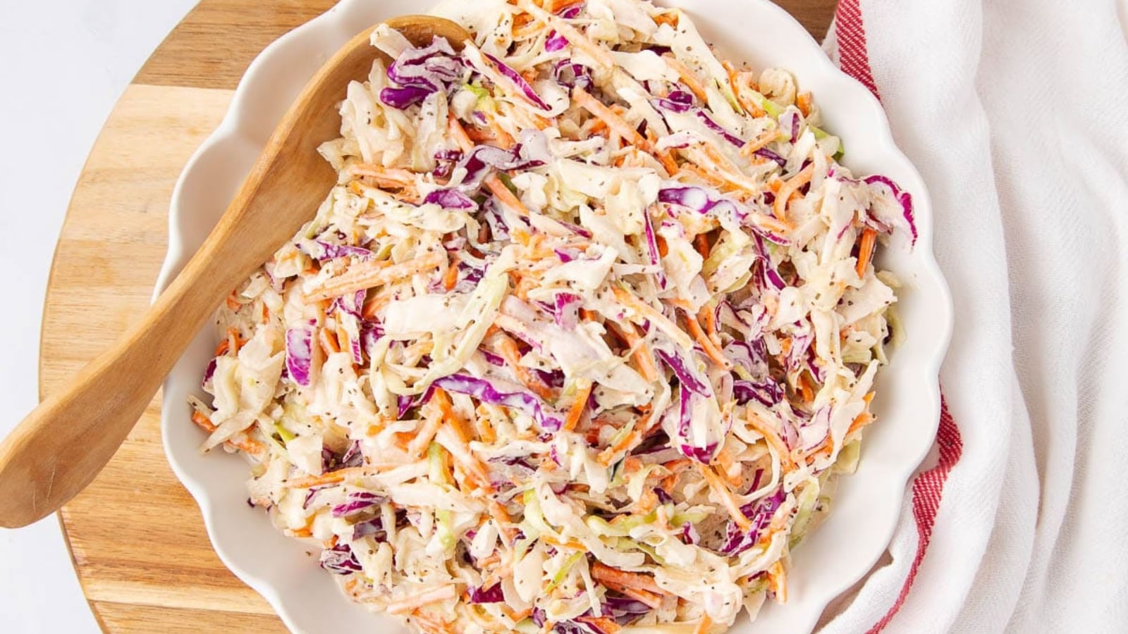 A bowl of coleslaw with shredded cabbage, carrots, and purple cabbage, mixed with creamy dressing, alongside a wooden spoon on a wooden board.