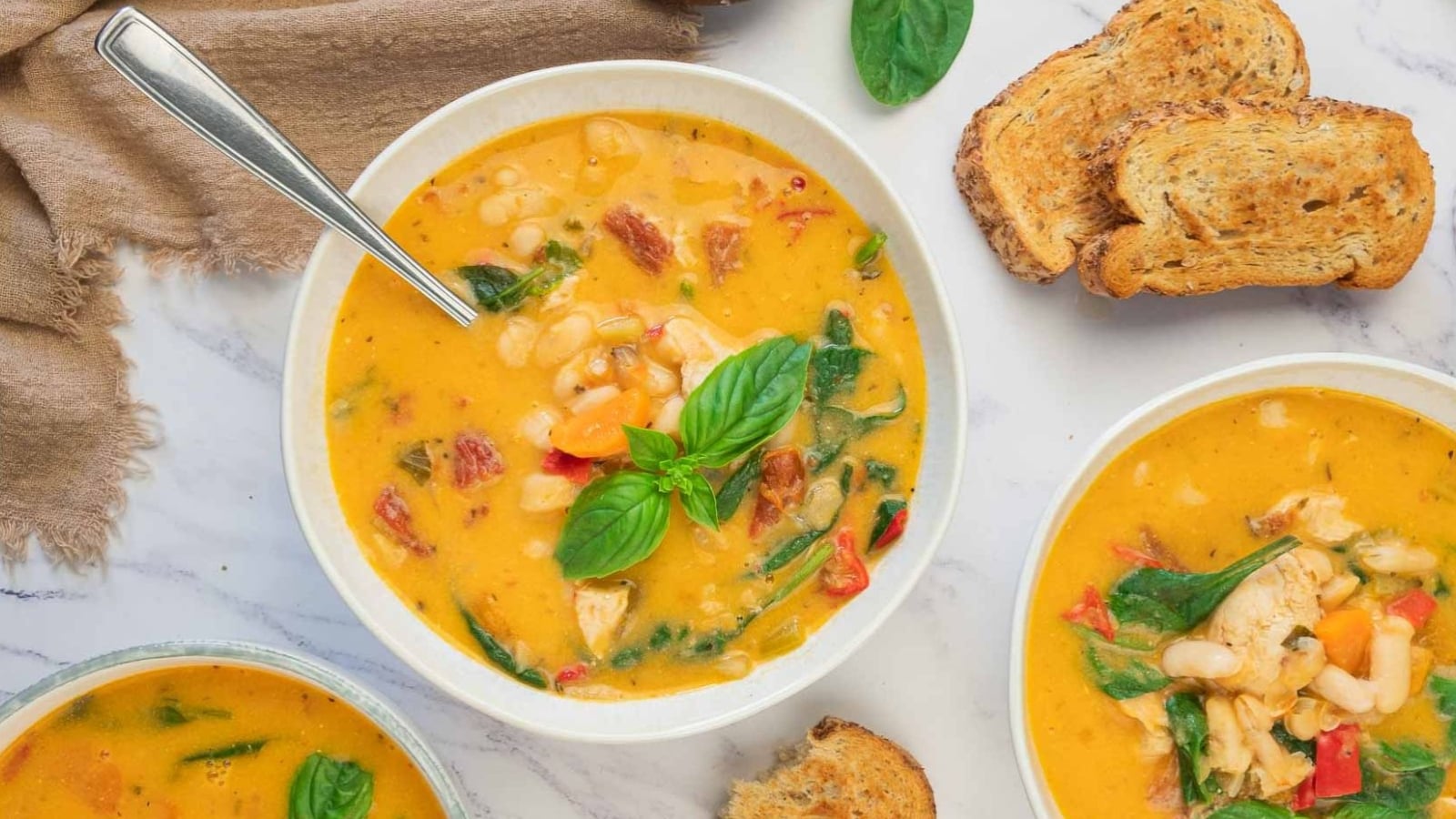 A bowl of creamy soup with beans, vegetables, and herbs, garnished with basil, served with toasted bread on a marble surface.