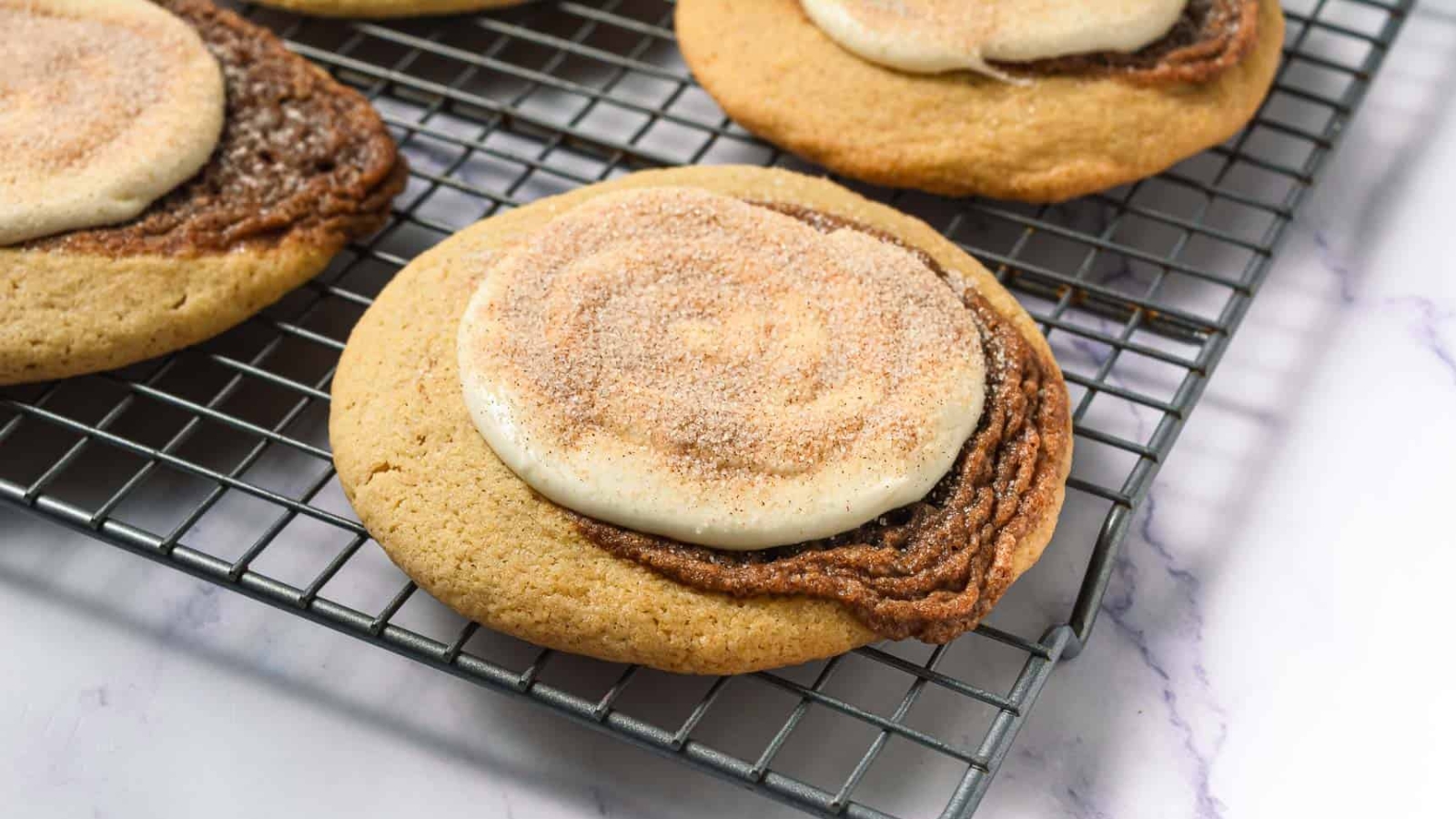 Four cinnamon swirl cookies on a white plate with a glass of milk and cinnamon sticks nearby.