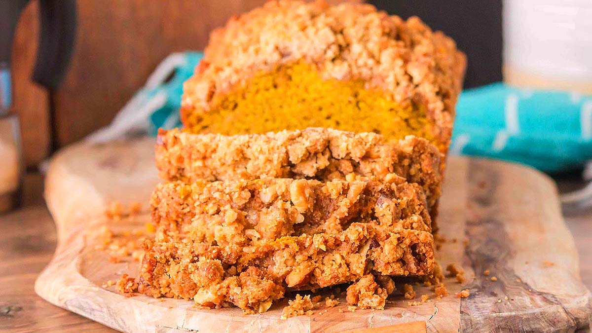 A loaf of crumb-topped pumpkin bread is sliced on a wooden cutting board, showing its moist orange interior and crumbly topping.