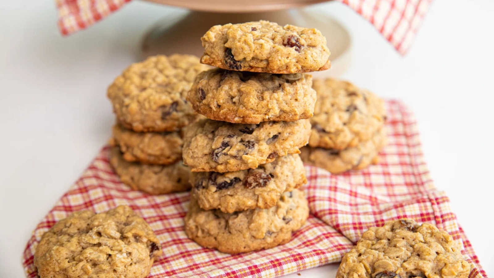 A stack of oatmeal raisin cookies sits on a red and white checkered cloth, with more cookies and a plate in the background.