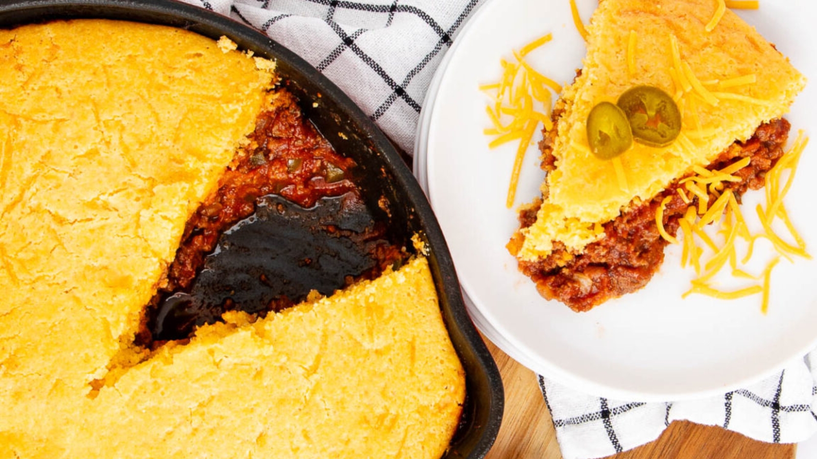 A baking dish of tamale pie with one slice removed, served on a plate with shredded cheese and jalape&ntilde;o slices.