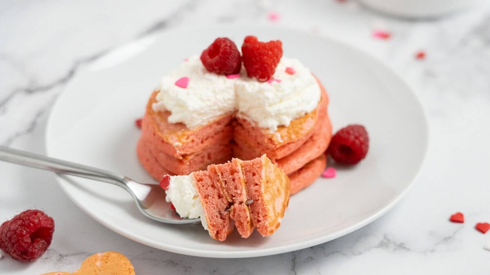 A stack of pink pancakes with whipped cream and raspberries, with a bite on a fork, on a white plate.