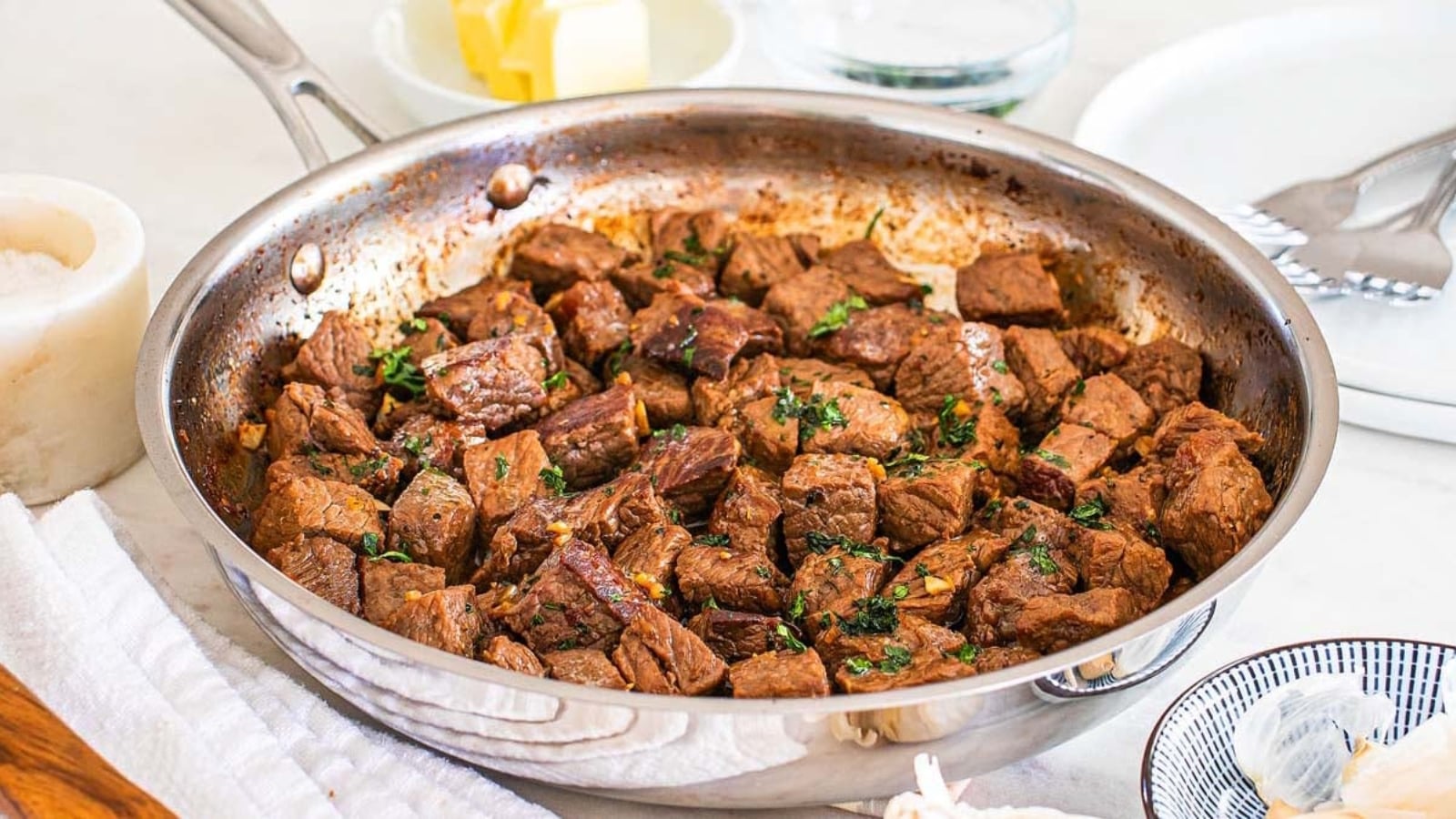 Chunks of seared steak with herbs in a stainless steel skillet, with butter and other ingredients in the background on a white countertop.