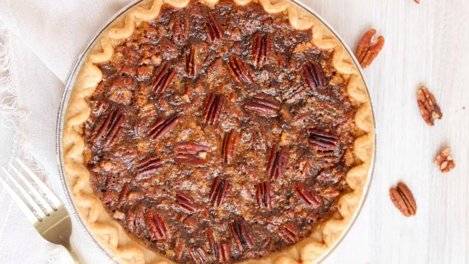 A whole pecan pie in a metal pie dish, viewed from above, with a golden crust and pecan halves on top. A fork and loose pecans are nearby on the table.