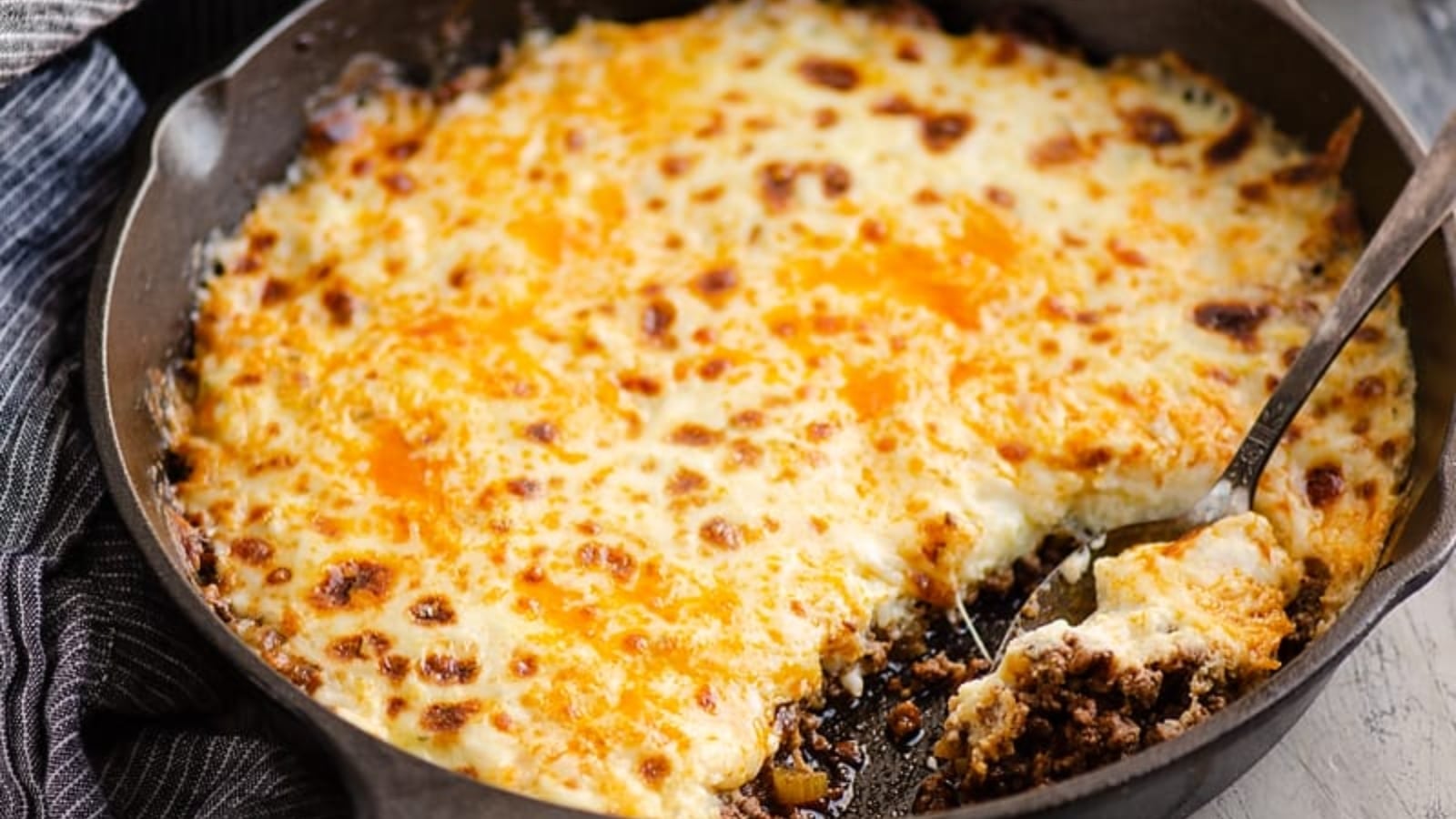 A skillet filled with baked ground meat and vegetables topped with a golden, melted cheese layer; a serving has been scooped out with a metal spatula.