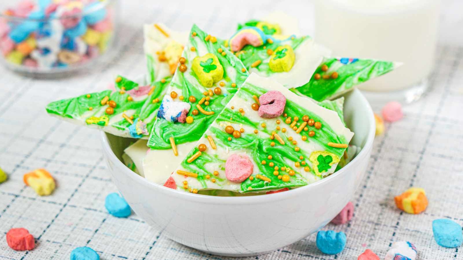 A bowl of festive leprechaun bark with marshmallows and sprinkles. A glass of milk and a bowl of Lucky Charms marshmallows are in the background.