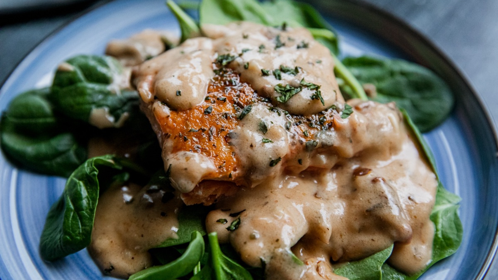 A piece of cooked fish covered in creamy sauce sits on a bed of fresh spinach leaves on a blue plate.