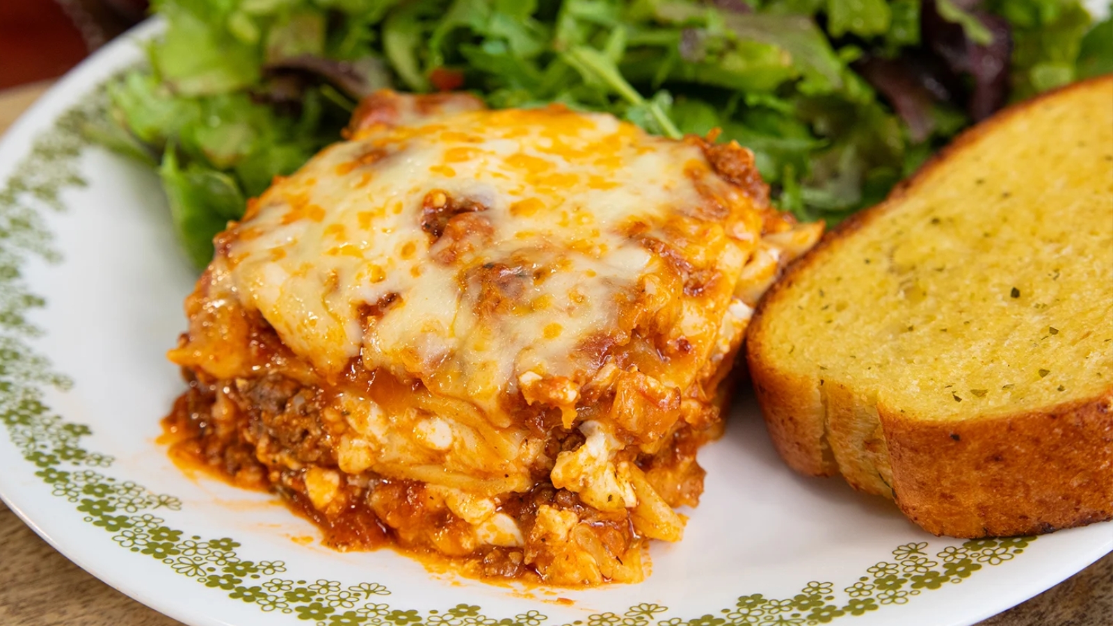 A serving of cheesy lasagna with meat sauce on a plate, accompanied by a slice of garlic bread and a green salad.