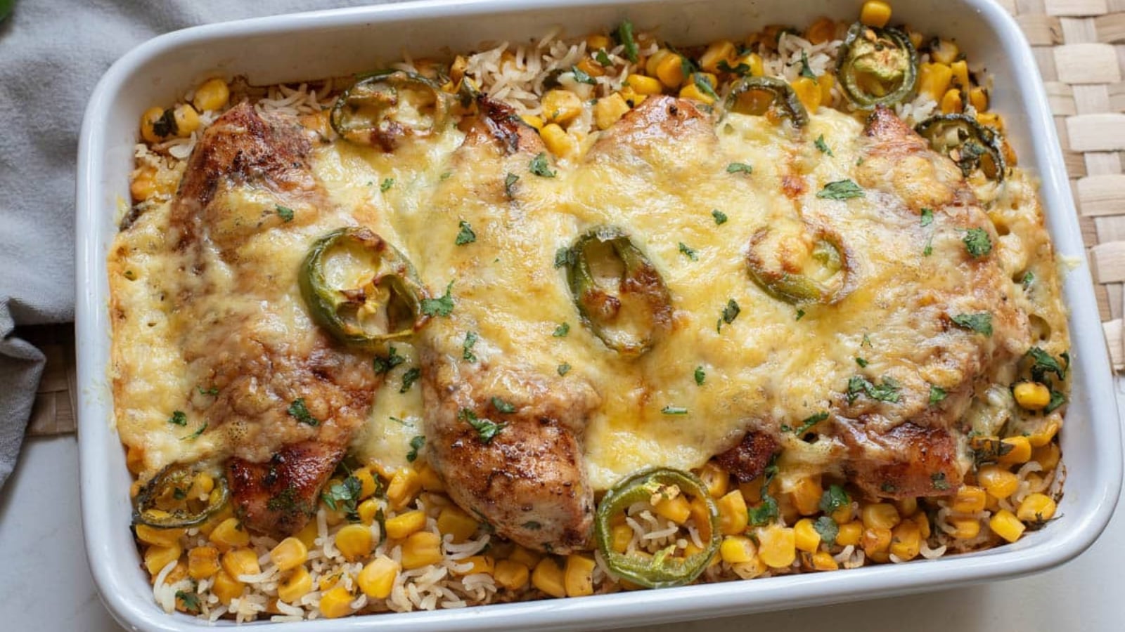 A baked casserole with chicken breasts topped with melted cheese and sliced jalape&ntilde;os, resting on a bed of rice and corn in a white dish.