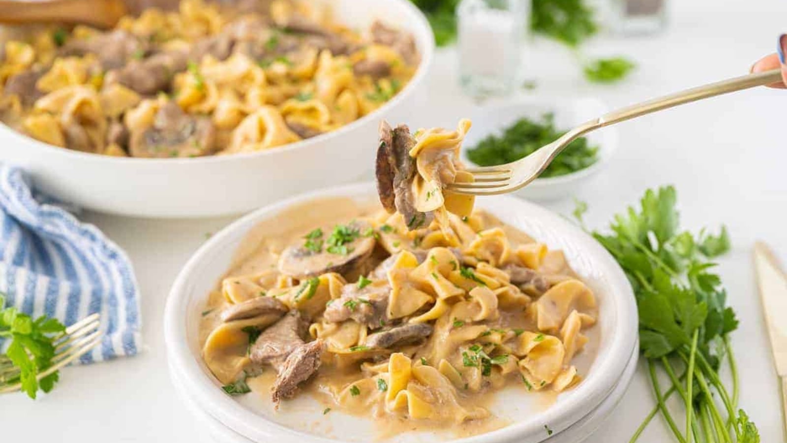 A plate of beef stroganoff with egg noodles and a creamy sauce is served, with a fork lifting a bite. Fresh parsley and a dish of stroganoff are visible in the background.