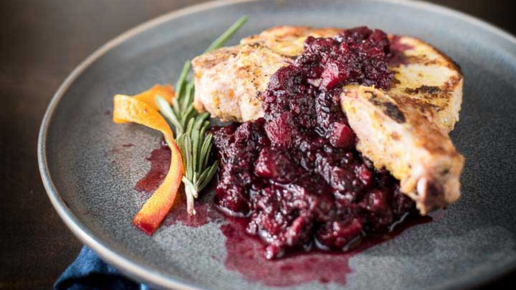 A seared pork chop topped with chunky cranberry sauce, garnished with a sprig of rosemary and orange peel, served on a grey plate.