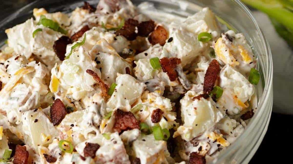 A close-up of a bowl of creamy potato salad with chopped green onions, crispy bacon pieces, and black pepper on top.