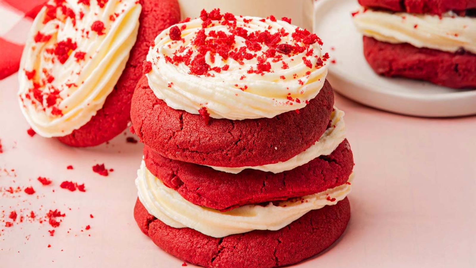 Two stacked red velvet sandwich cookies with cream cheese frosting filling, topped with red crumbs, sit on a pink surface beside two more cookies and a glass of milk.