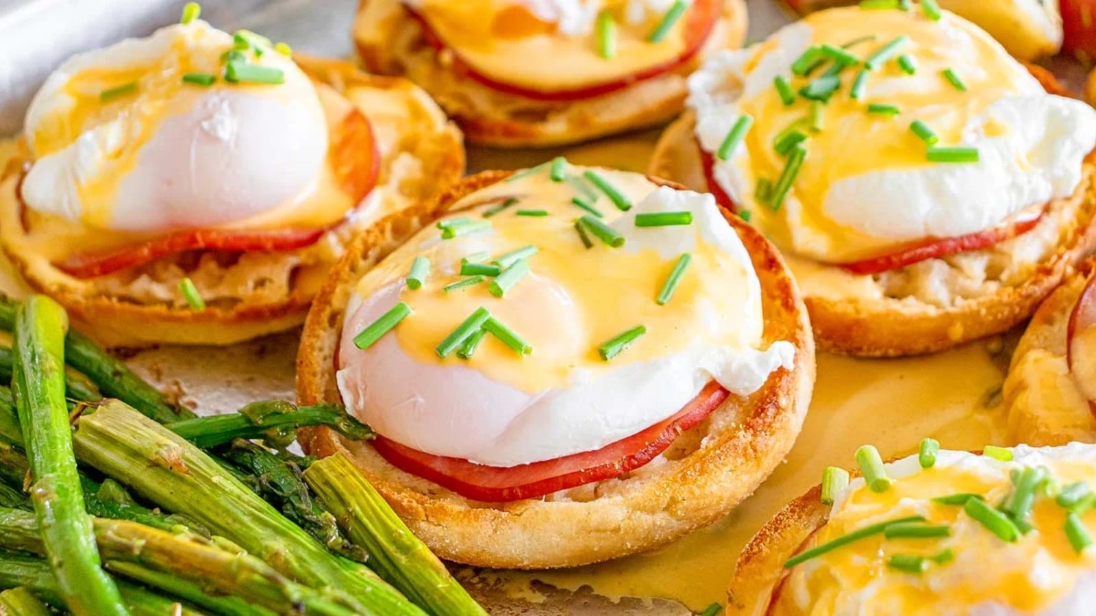 English muffins topped with ham, poached eggs, hollandaise sauce, and chopped chives, served with a side of cooked asparagus.