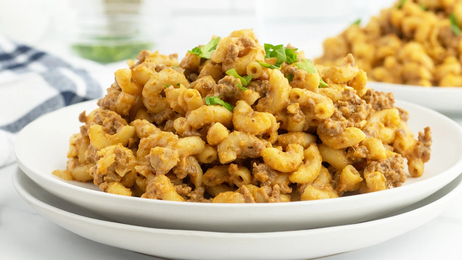 A plate of macaroni pasta mixed with ground meat and garnished with chopped herbs, served on a white dish.