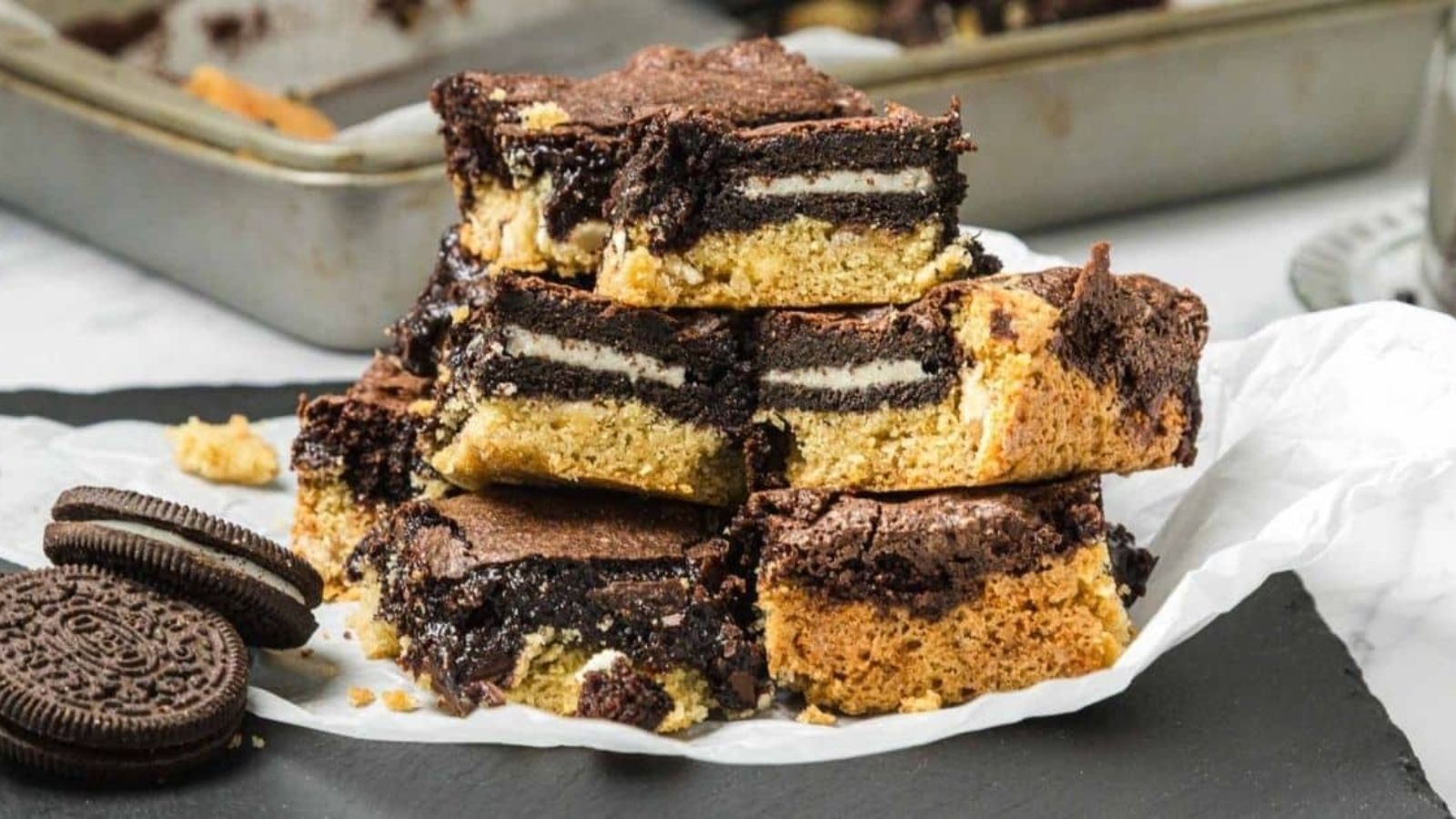 A stack of layered cookie and brownie bars with Oreo cookies inside, displayed on parchment paper.