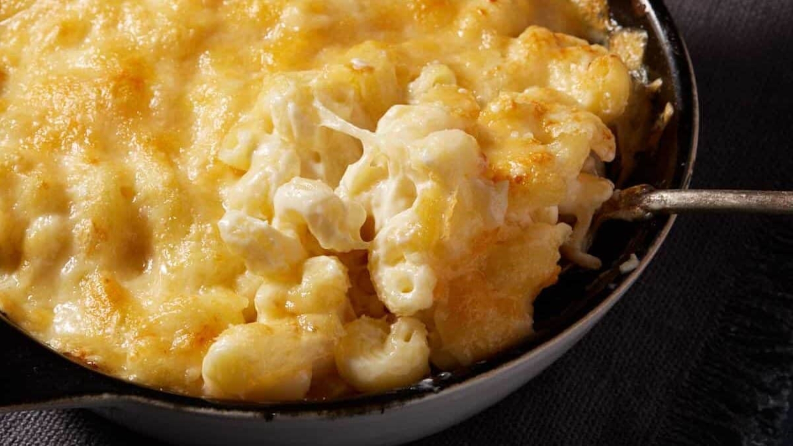 A close-up of baked macaroni and cheese in a round dish, with a spoon lifting a serving to show the creamy, melted cheese topping.