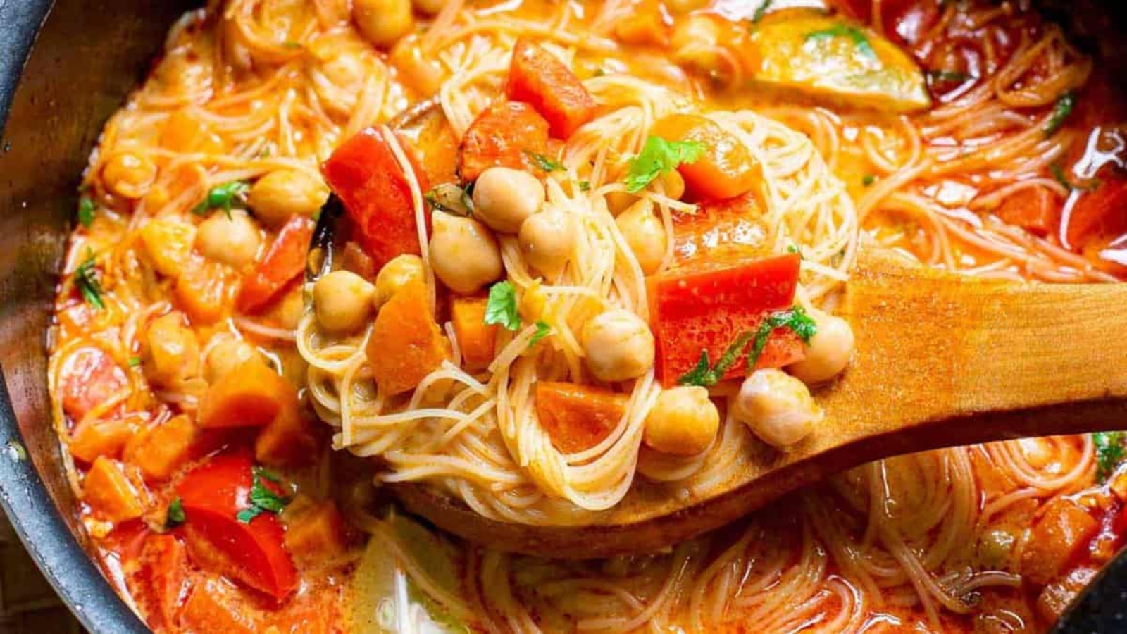 A close-up of a wooden spoon lifting vegetable and chickpea spaghetti in tomato sauce from a pot.