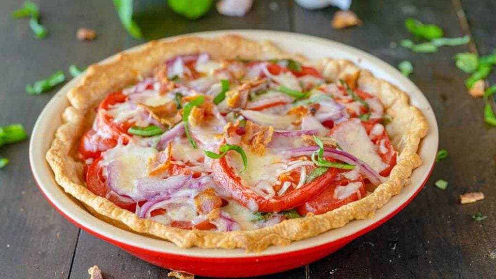A baked pie with a golden crust, topped with sliced tomatoes, red onions, green herbs, cheese, and bacon pieces, displayed on a wooden table.