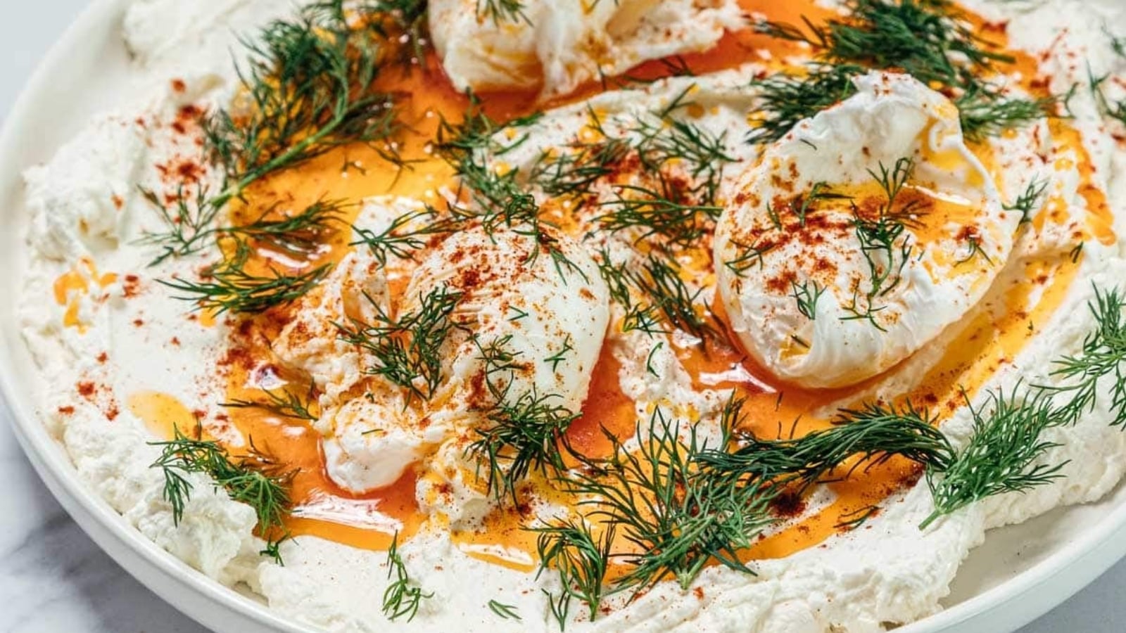 Creamy yogurt topped with poached eggs, paprika oil, and fresh dill on a white plate.