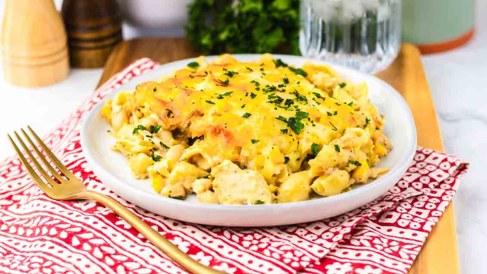A plate of cheesy pasta casserole garnished with chopped parsley, on a red-patterned napkin with a gold fork.