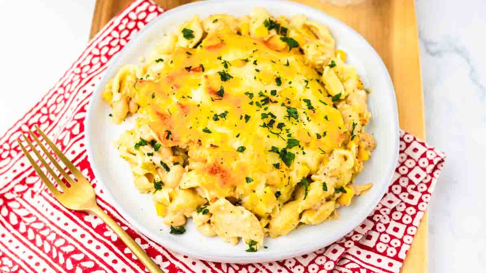 A plate of cheesy pasta casserole garnished with chopped parsley, on a red-patterned napkin with a gold fork.