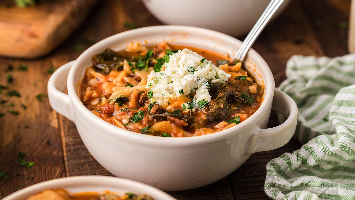 A bowl of tomato-based soup with rice, leafy greens, and a dollop of cheese on top, served with a spoon on a wooden table.