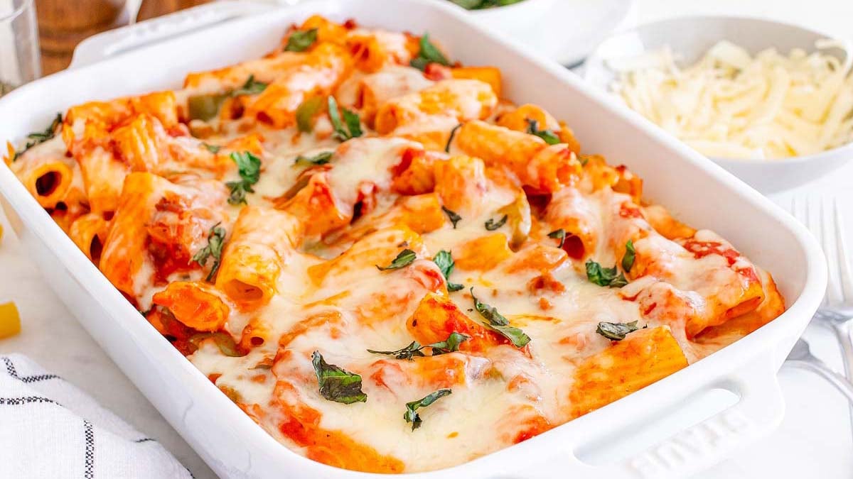 A white baking dish filled with baked rigatoni pasta, tomato sauce, melted cheese, and fresh basil leaves on top.