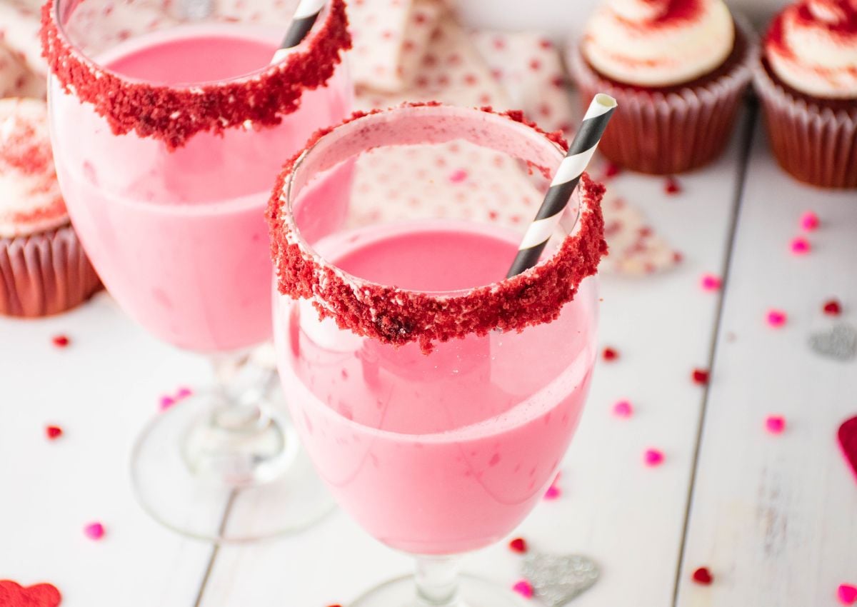 Two glasses of pink milkshake with black and white straws, rimmed with red crumbs, are on a white table next to decorated cupcakes and scattered confetti.